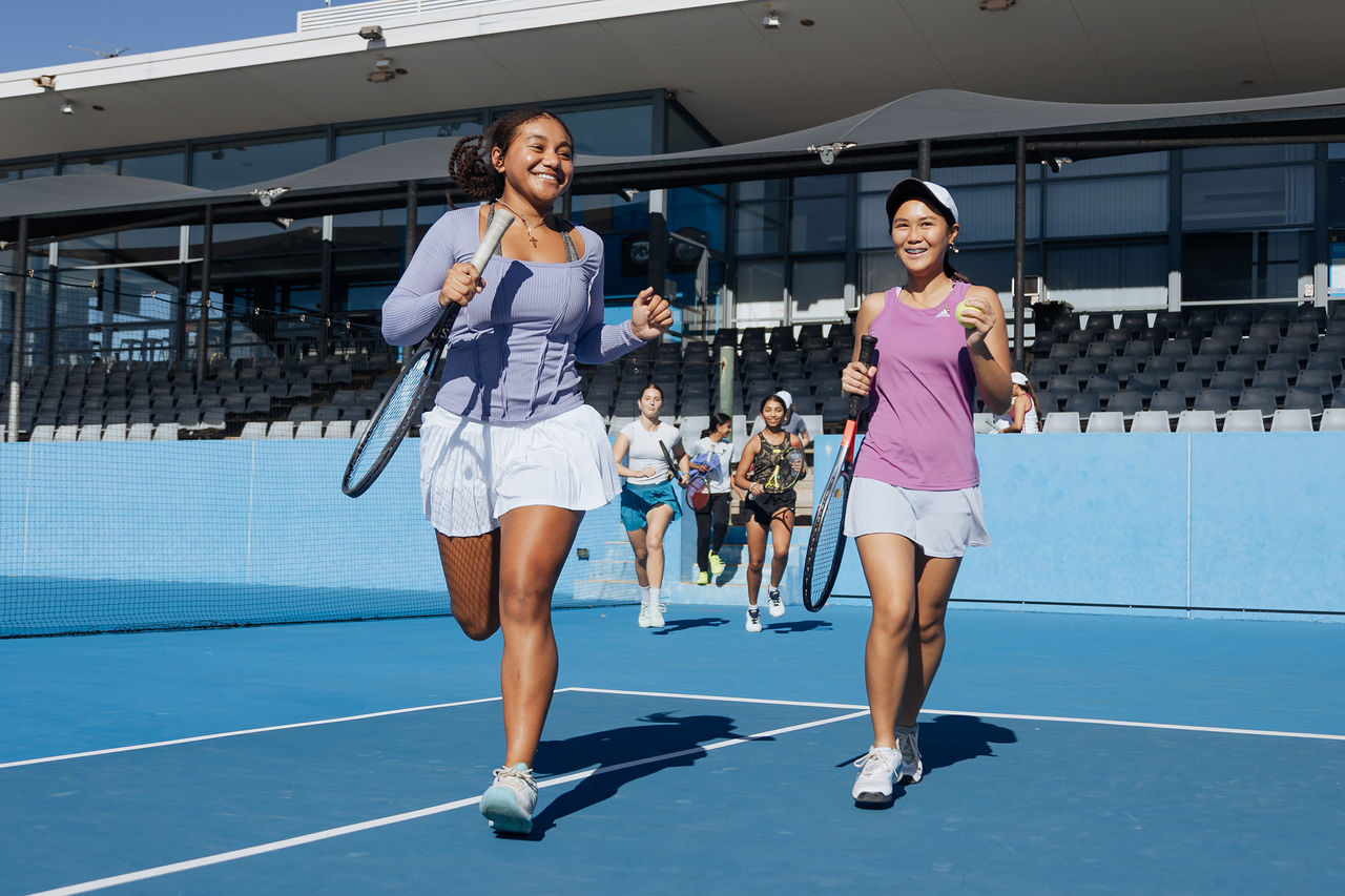 A group of girls clutching tennis racquets excitedly run onto court to play